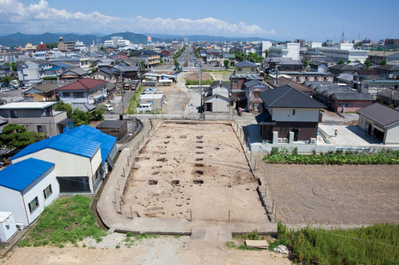 西から見た大型掘立柱建物跡、中央奥は県道、左上の白いビルは南国市役所、右上の白い建物は高知東工業高校の校舎(高知県南国市篠原)。公益財団法人 高知県立埋蔵文化財センター